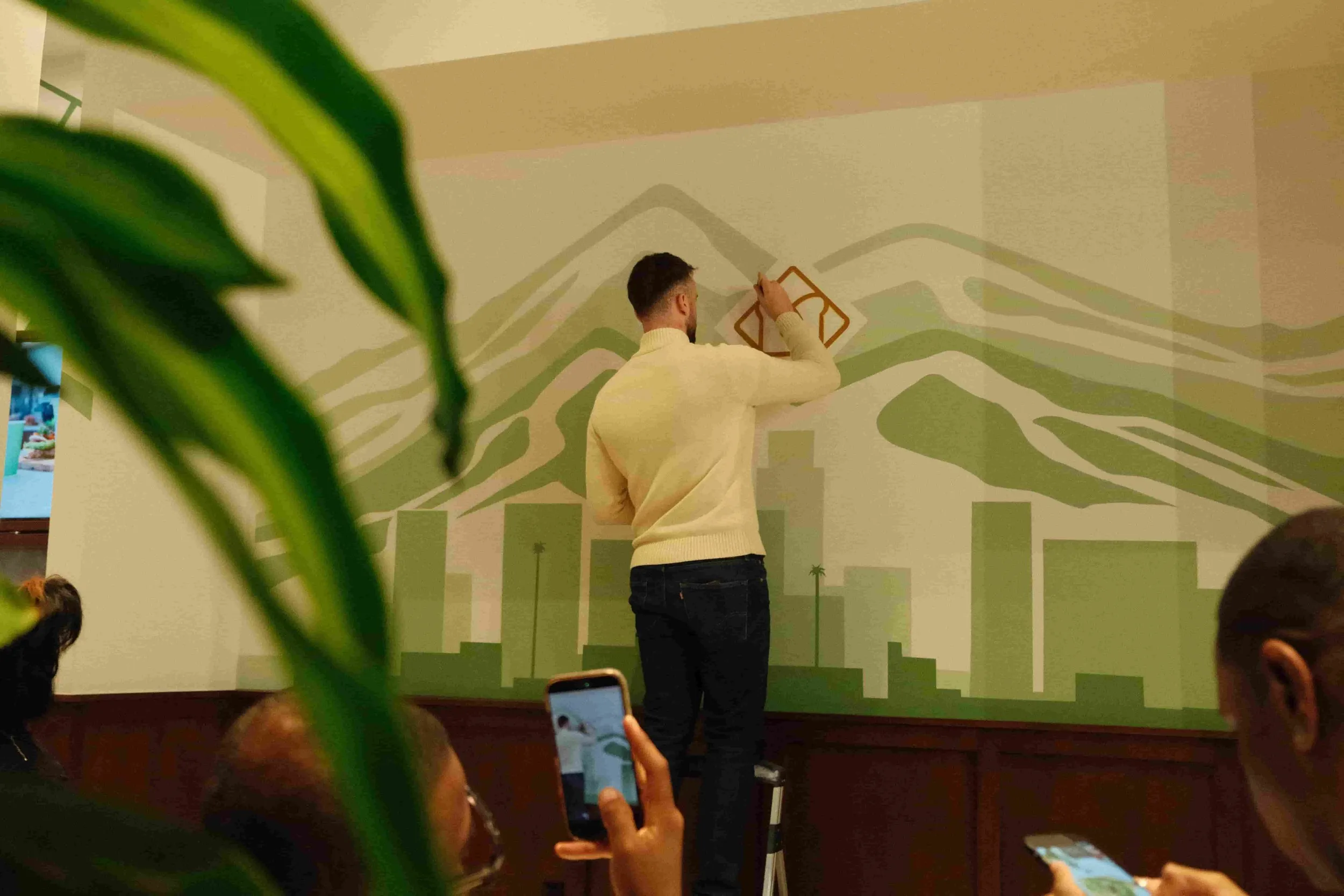 A man wearing a yellow sweater is drawing on a mural of a mountain landscape on a white wall. The scene includes stylized mountains and trees, with a green and beige color scheme. People in the foreground are taking photos and watching the artist.