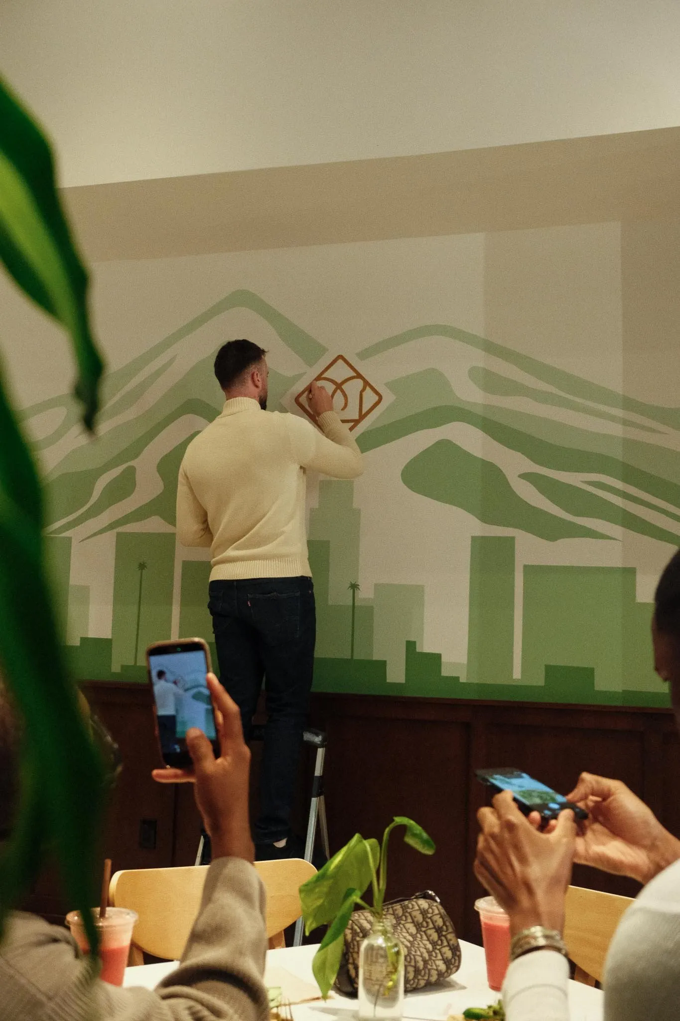 A man in a yellow sweater is drawing on a artwork or sign on a wall. Several people are sitting at a table with drinks, taking photos.