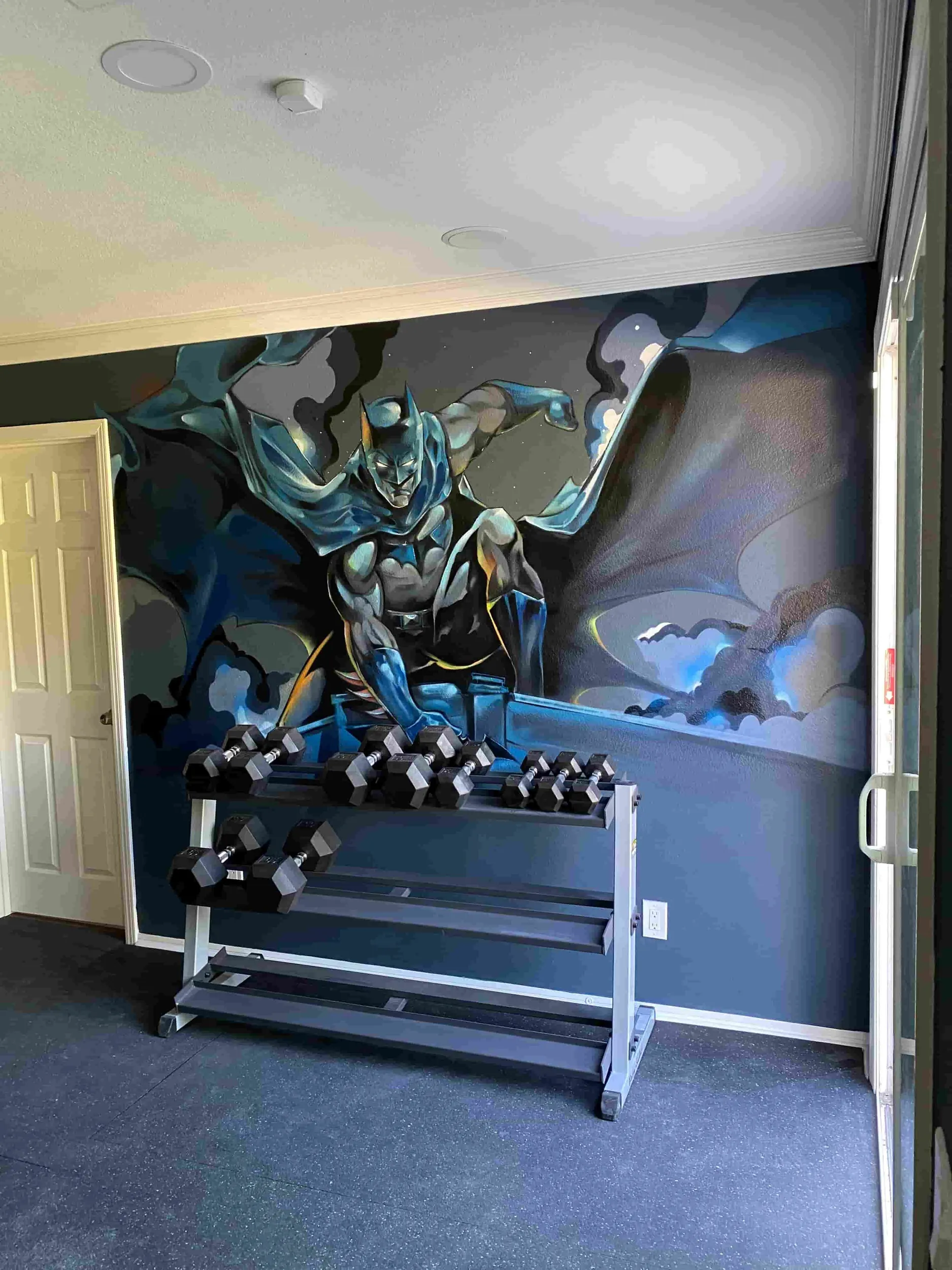 Gym with a large Batman mural on the wall, black dumbbells on a white rack, and a gray and black floor. There is a white door partially open on the left and a white grab bar on the right.