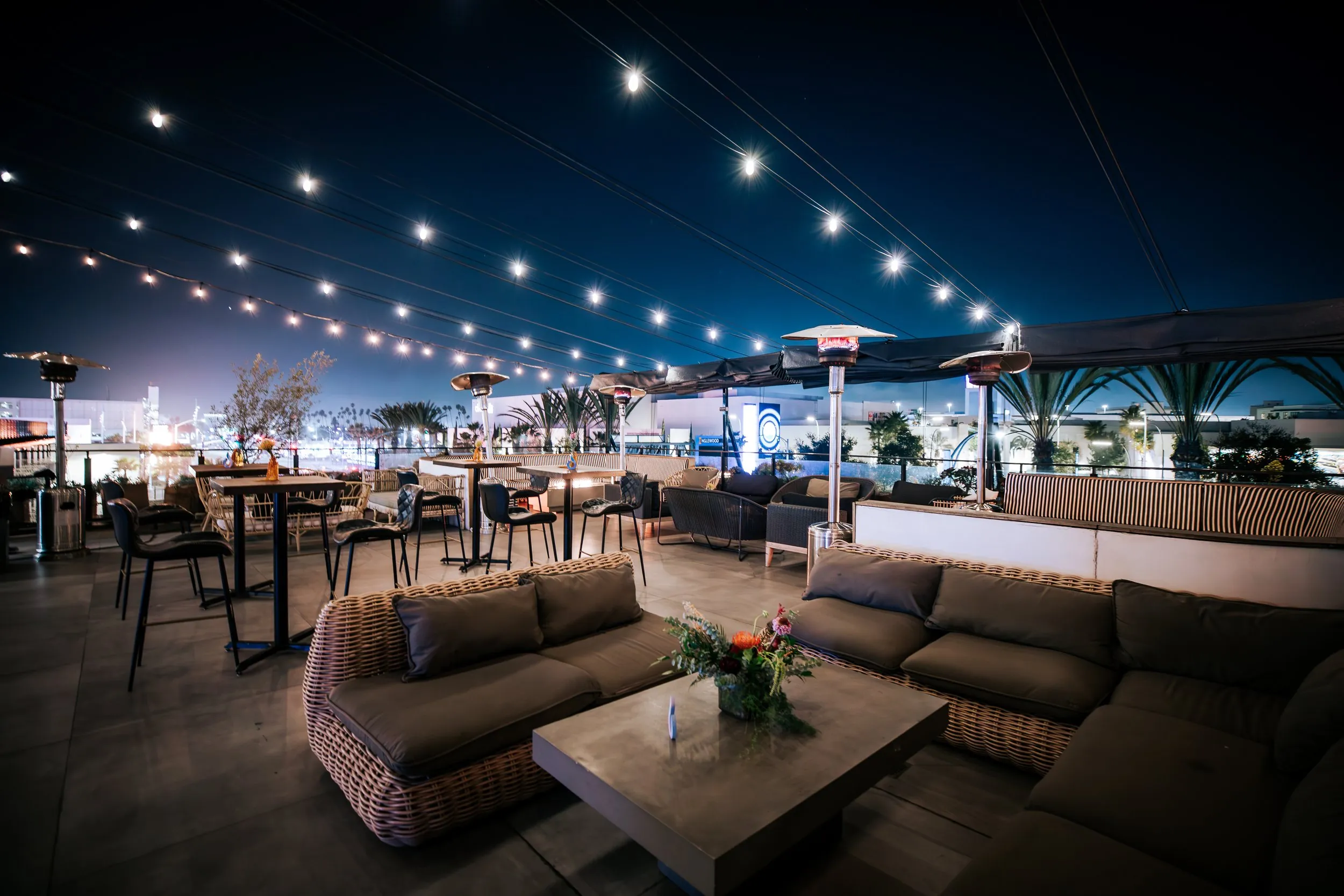 Nighttime rooftop patio with string lights, outdoor heaters, cushioned seating, tall tables, and cityscape views with palm trees in the background.