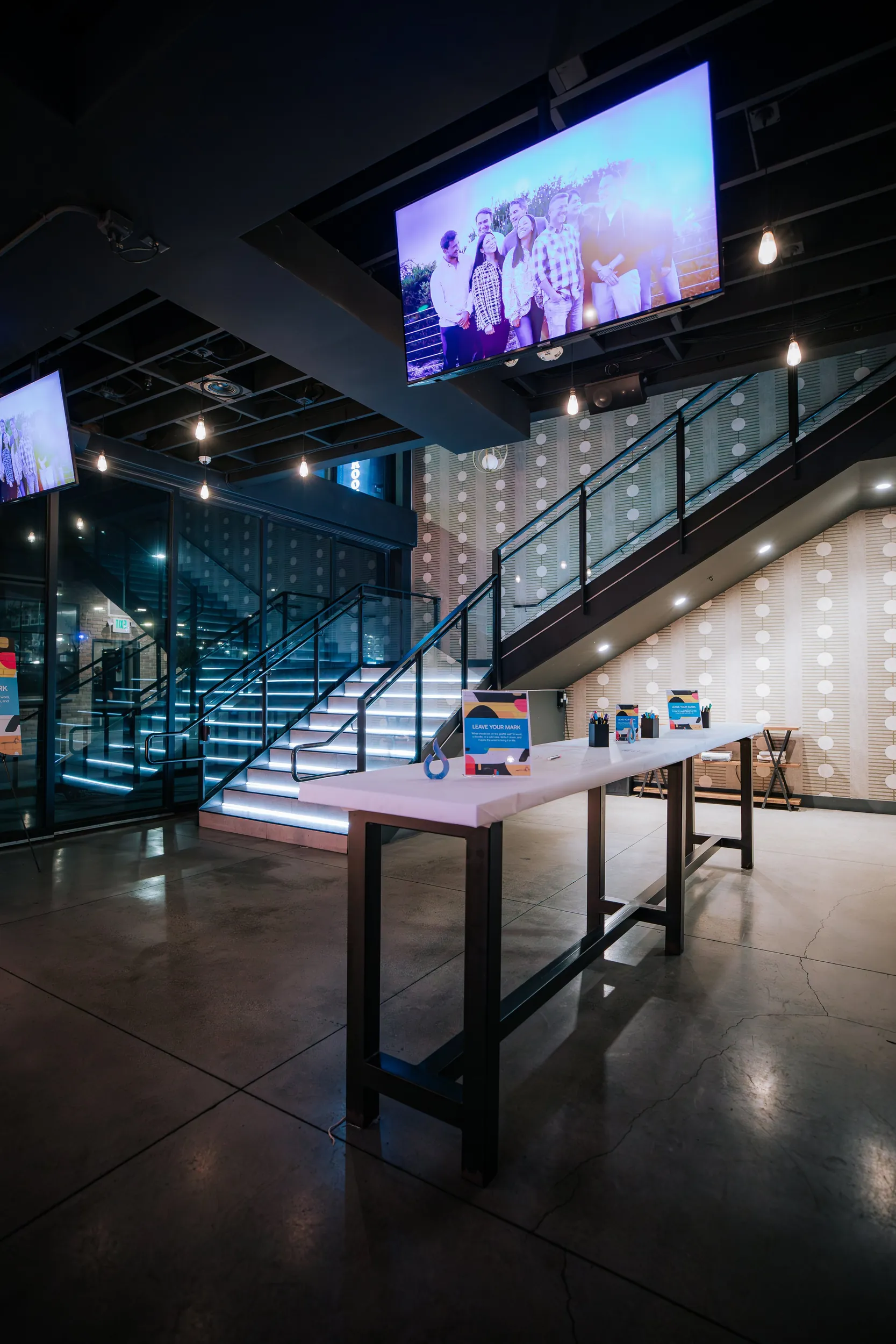 Interior of a modern event space or conference room with a staircase, large television screens displaying a group photo, and a long table with markers and informational signs.