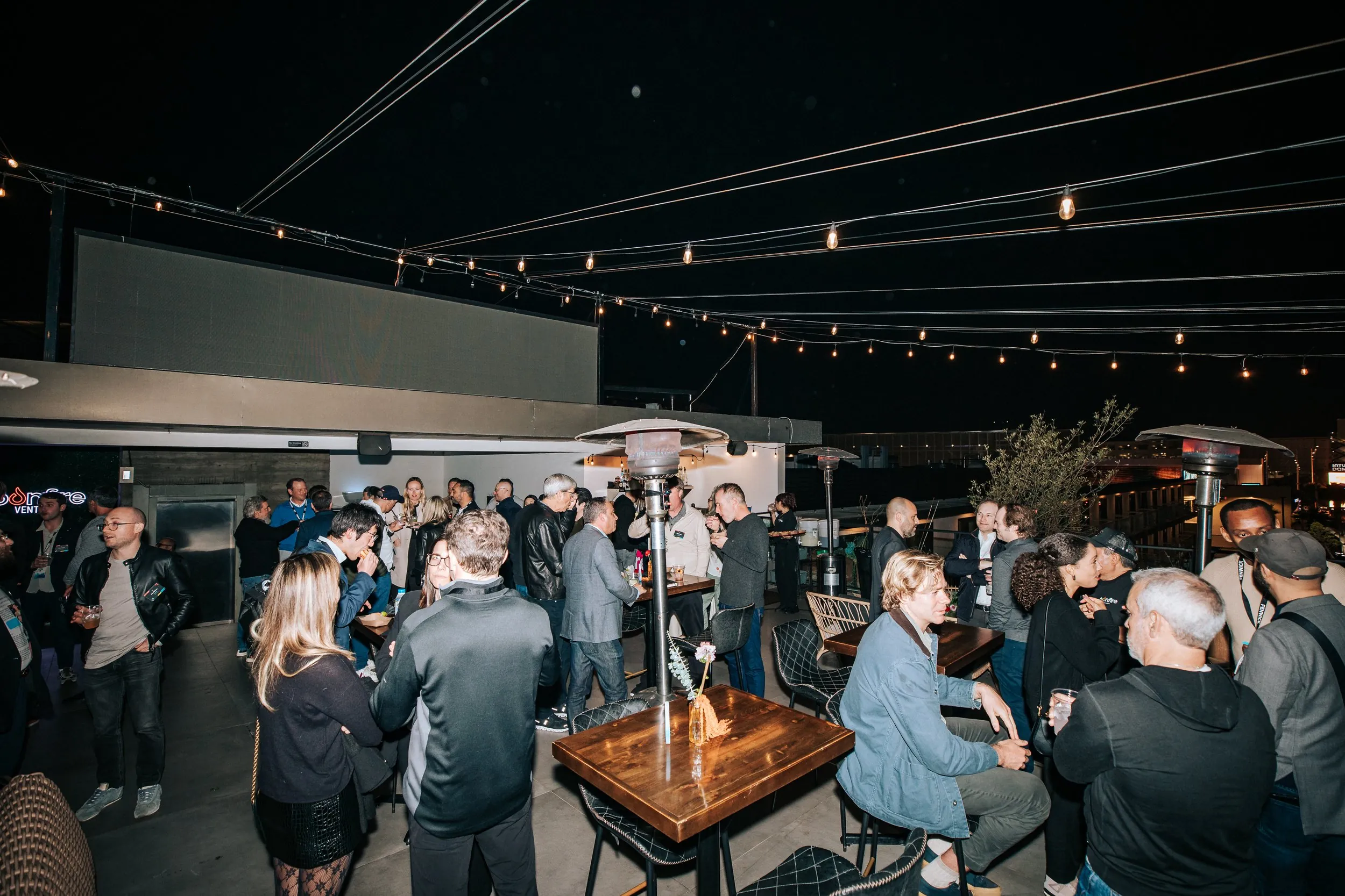 People socializing at an outdoor rooftop party at night with string lights overhead and patio heaters. Some are standing, others sitting at tables, holding drinks.