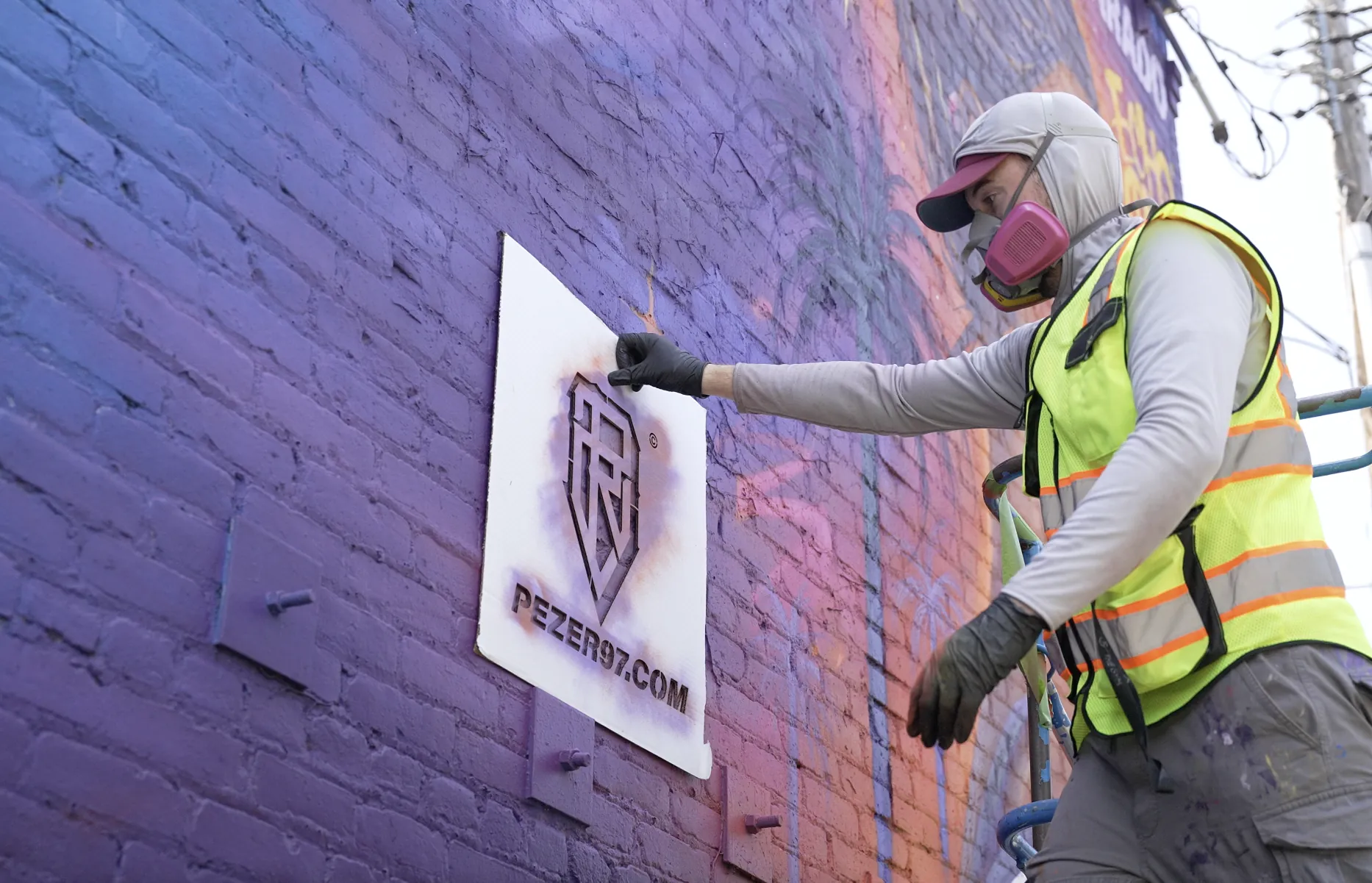 A person in a high-visibility safety vest, wearing a gray hoodie, pink face mask, and gloves, is installing or adjusting a sign on a purple mural art wall. The sign displays a logo and the website pezer97.com.
