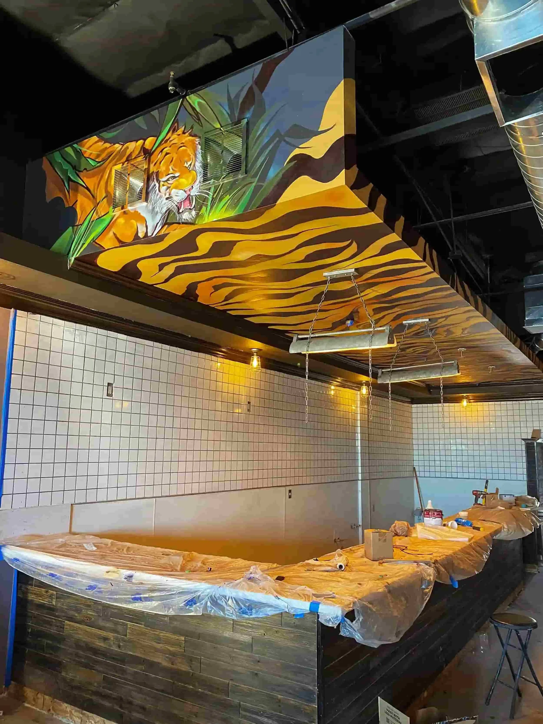 Interior of a restaurant or bar under construction or renovation, with a ceiling mural of a jungle scene featuring a tiger among lush green foliage, and tiger stripe patterns on the lower part of the ceiling. Construction materials and tools are visible on a covered bar counter and in the surrounding area.