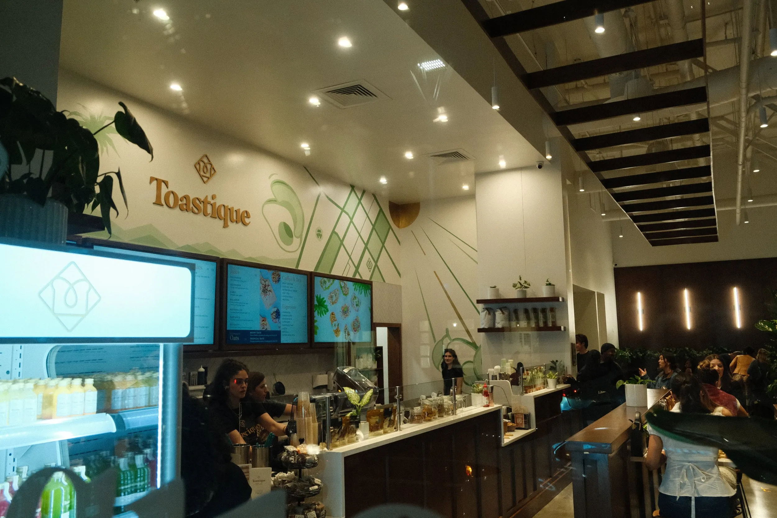 Interior of a cafe named Toastique with customers waiting in line and staff behind the counter serving drinks and food. The decor includes wall art and plants, with a modern ceiling design featuring black beams and spotlights.