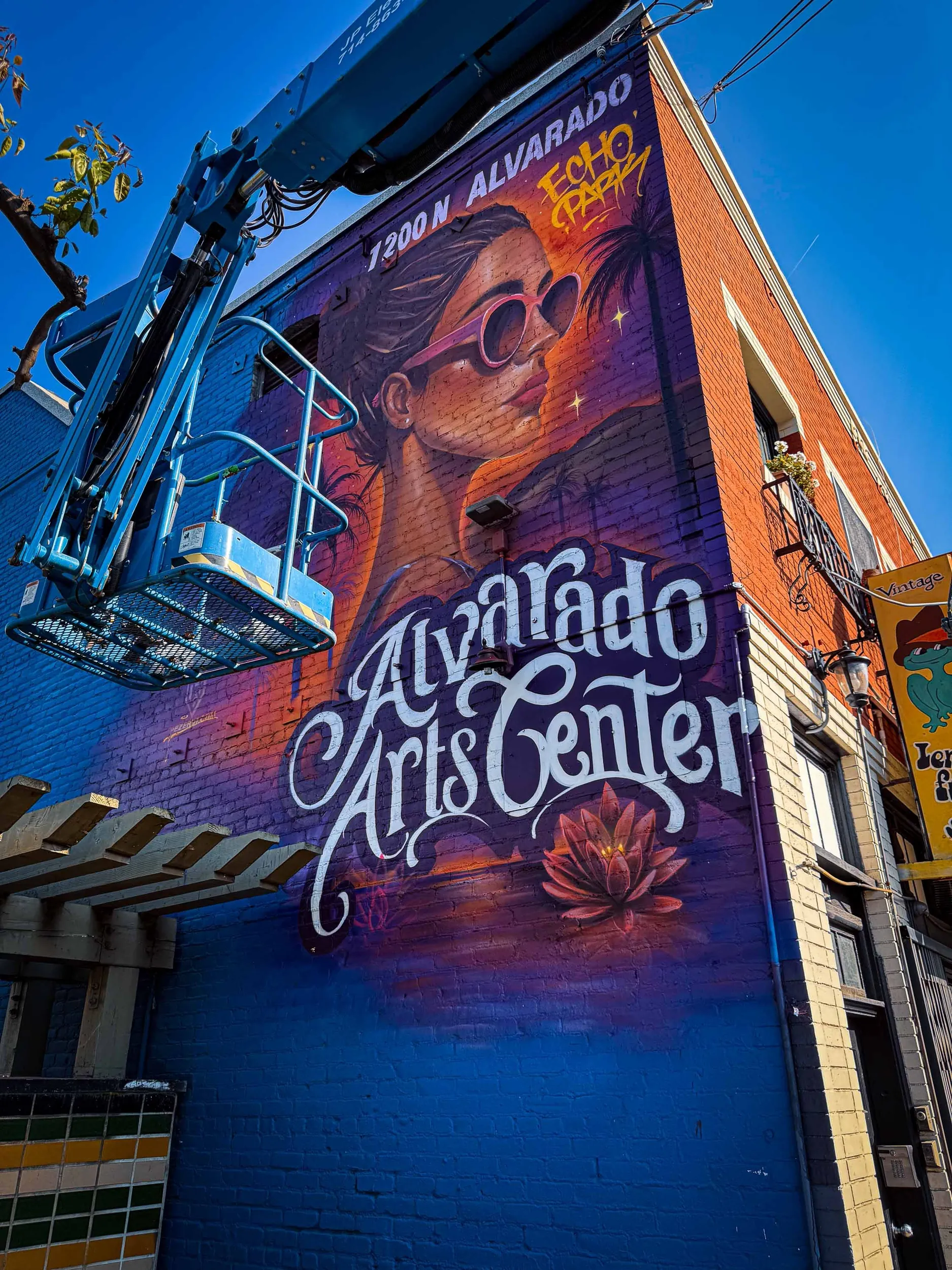 Colorful mural on the side of a building featuring a woman with sunglasses, a palm tree, a lotus flower, and the words