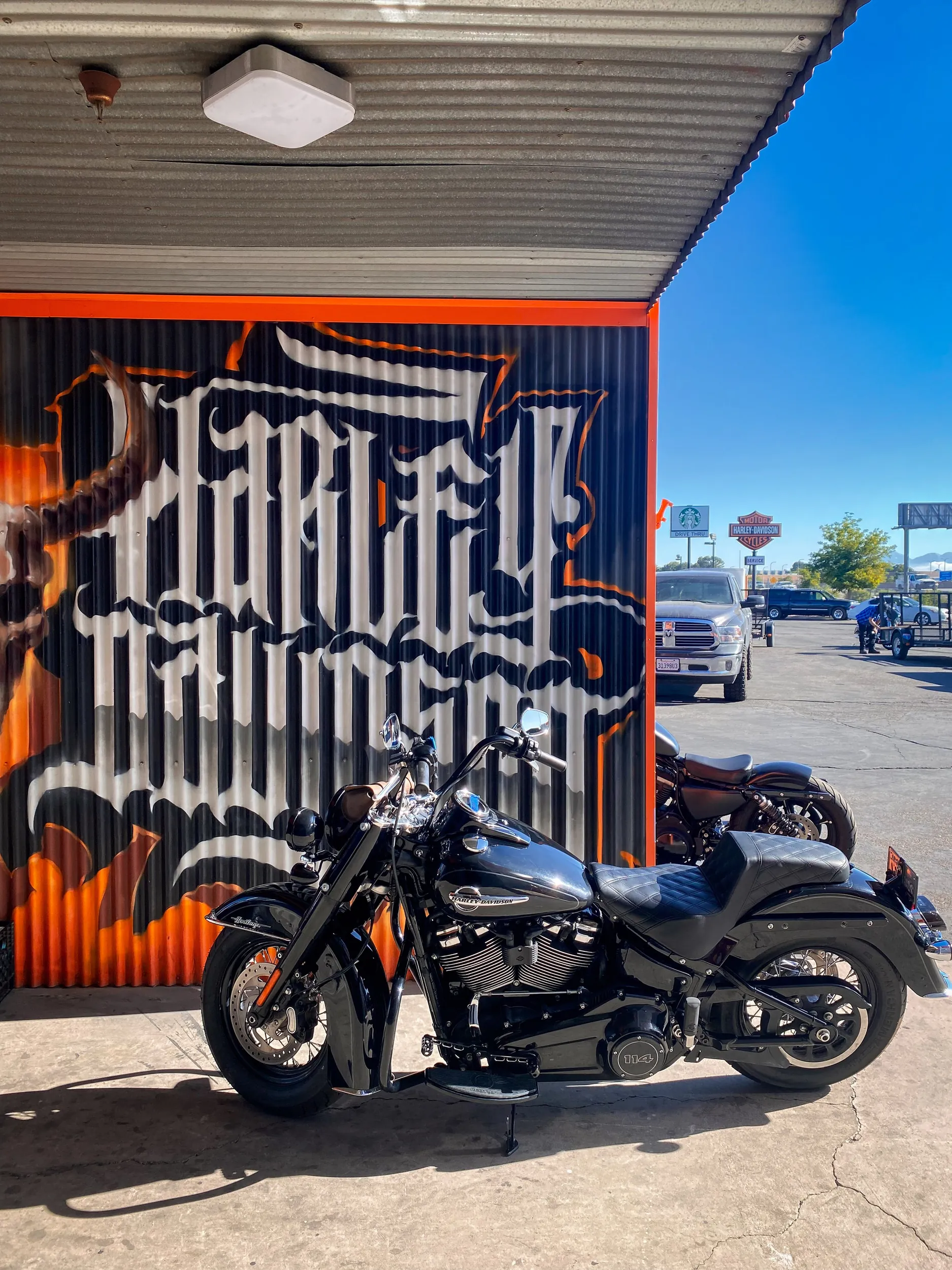 A black Harley Davidson motorcycle parked outside a building with orange and black graffiti on the wall, under a corrugated metal roof. In the background, there are various vehicles, including trucks and a golf cart, and signs for Harley Davidson and other businesses.