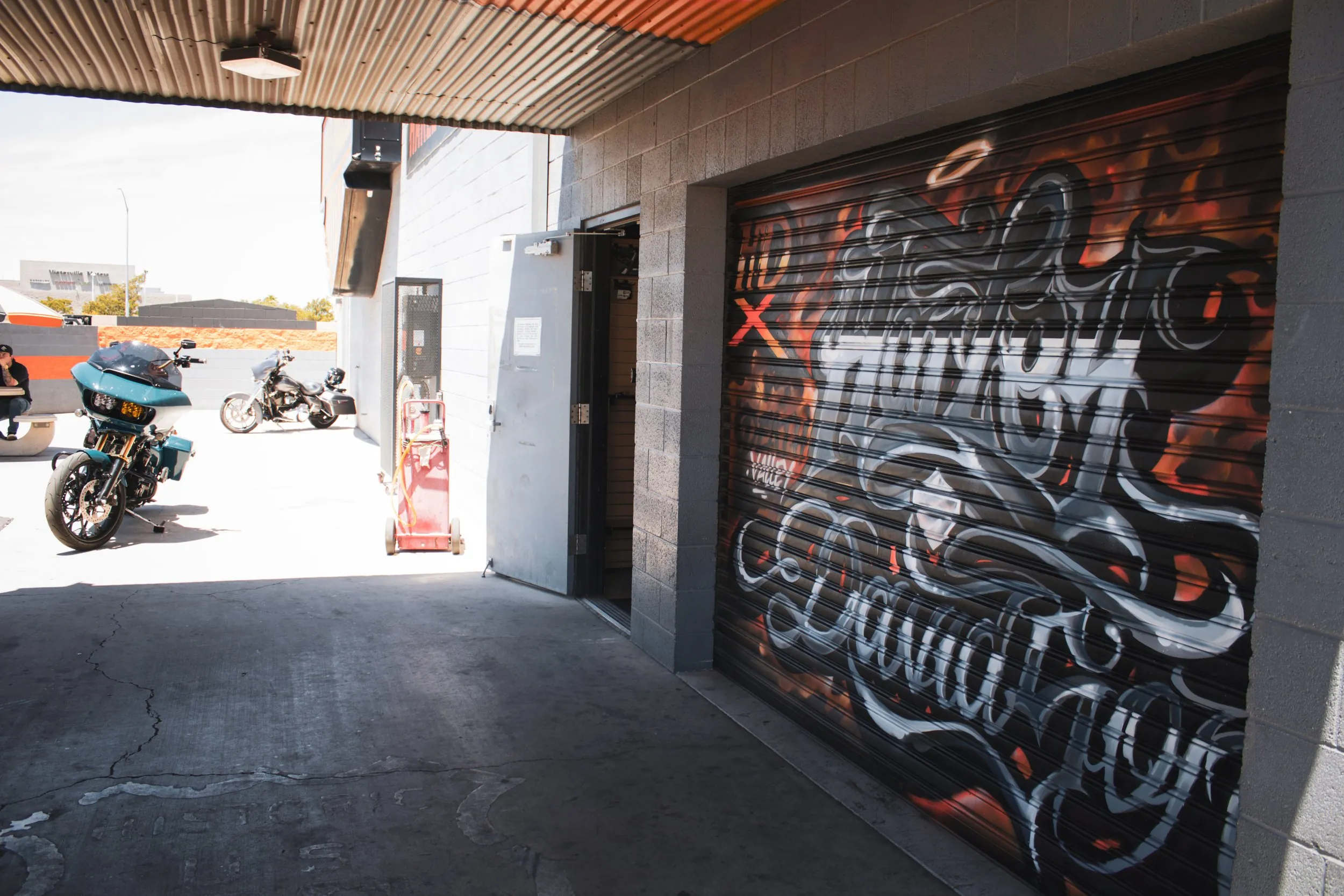 A garage with a mural on the rolled-up garage door, depicting stylized graffiti art with swirling patterns in black and white with red accents. Outside, there are three motorcycles parked, and a person is sitting on a bench on the left side.