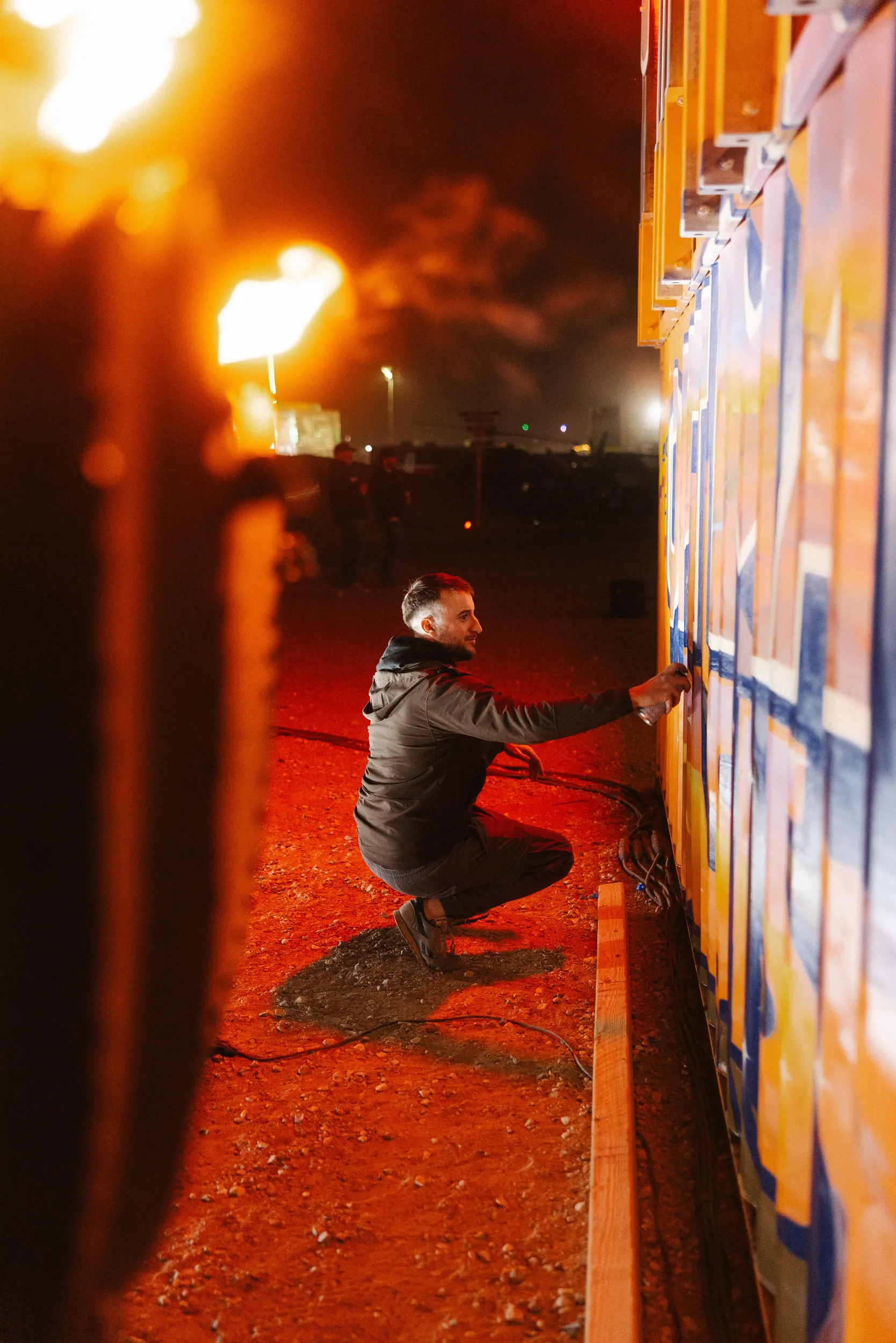 A man kneeling on the ground, drawing or writing on a colorful wall mural at night, with bright lights and smoke in the background.