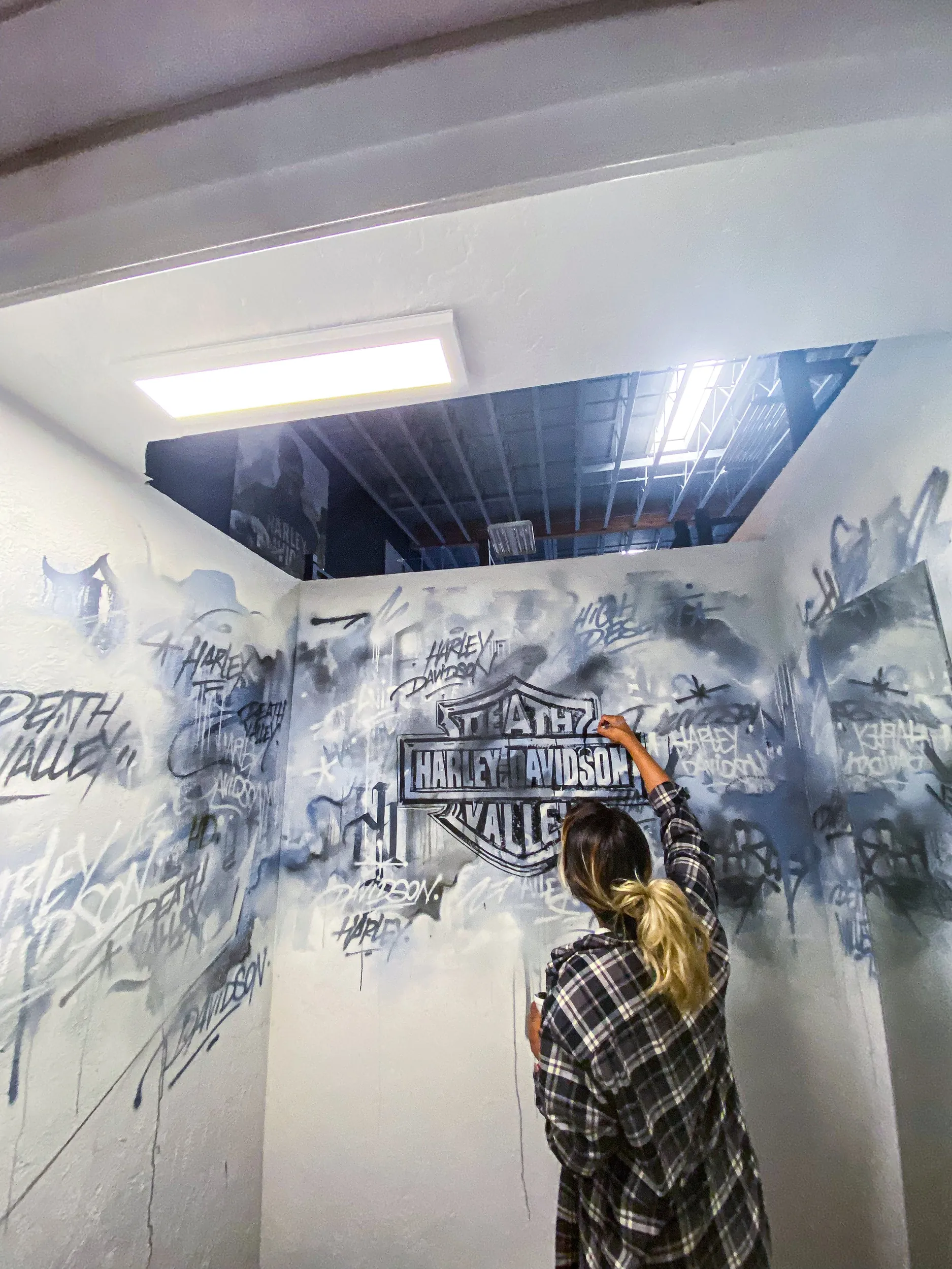 A person with long hair tied in a ponytail, wearing a black and white plaid shirt, spray-painting a Harley Davidson logo on a white wall that also has multiple graffiti tags and texts, in an indoor space with a ceiling opening and a skylight.