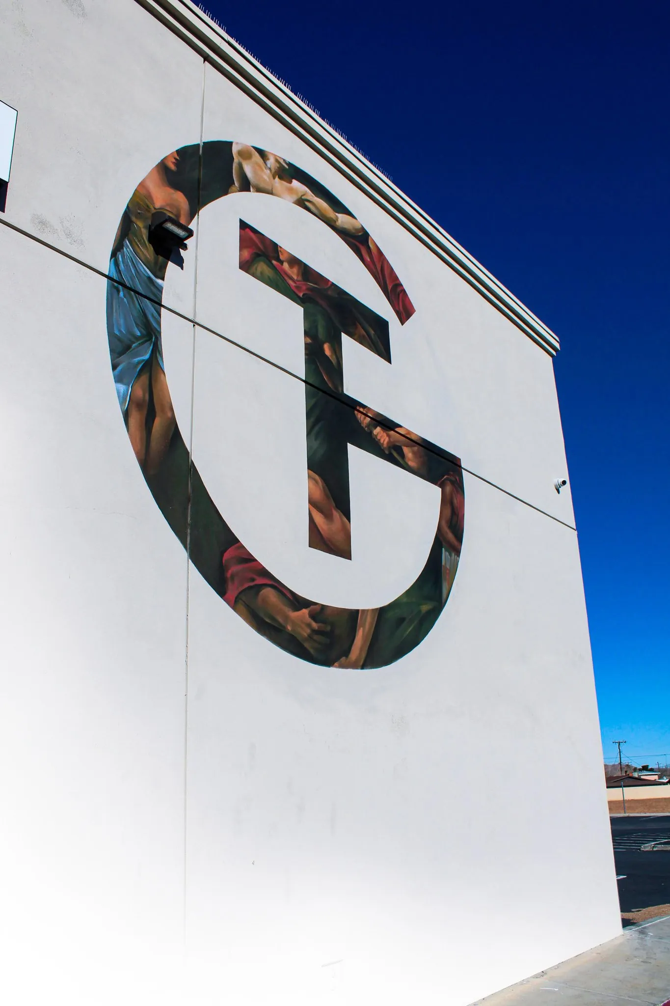 The image shows a large white building with a mural of a basketball player on the side. The mural is stylized with colorful and abstract elements making up the figure of the player in a dynamic pose. The sky is clear with a deep blue color, and there is some parking lot visible at the bottom right corner.
