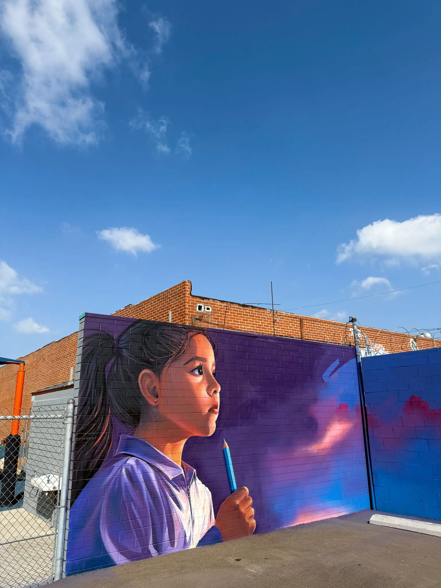 A colorful mural of a young woman holding a blue marker in front of a blue and purple background, painted on a brick wall.