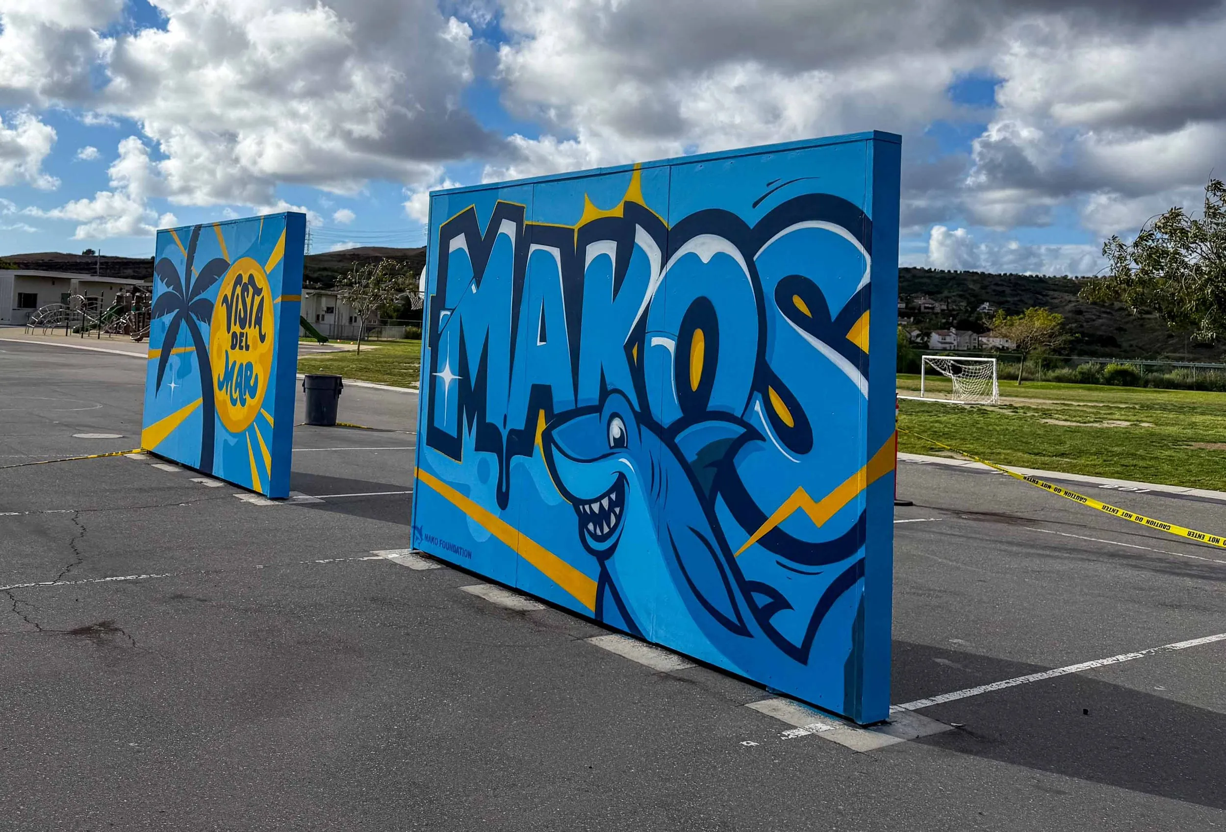Graffiti art on three large panels in a park, with a blue background, yellow sunburst, palm trees, and a stylized shark face with sharp teeth. The panels contain words and artistic designs, with a yellow caution tape outlining the area.